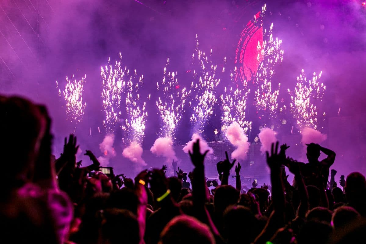 Mulțime de oameni dansând la un concert, sub lumini mov și artificii de scenă